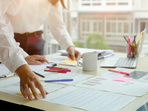 Two people from mid-chest down, standing at a desk reviewing various charts and documents. The table has a laptop, coffee cup, and other office supplies including sticky notes.