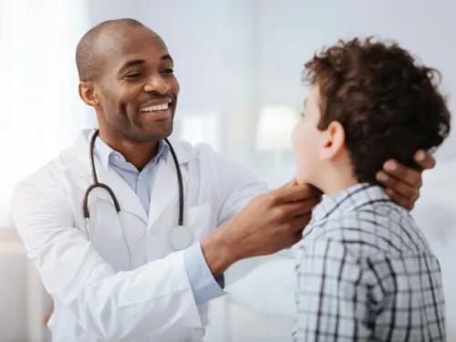 A smiling doctor in a white coat with a stethoscope around his neck is examining a child with a plaid shirt who is also smiling.