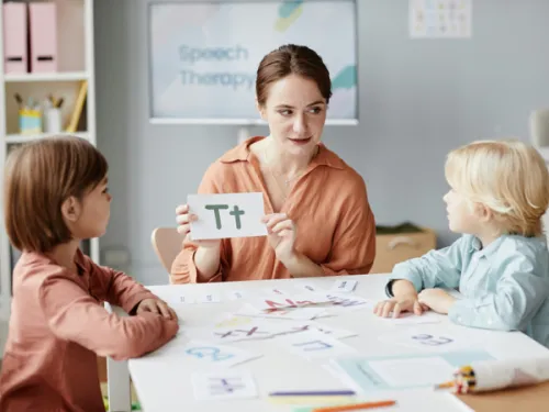 A female teacher sitting at a table with two young students under the age of 10, who is holding a card that has an uppercase and lowercase letter T.